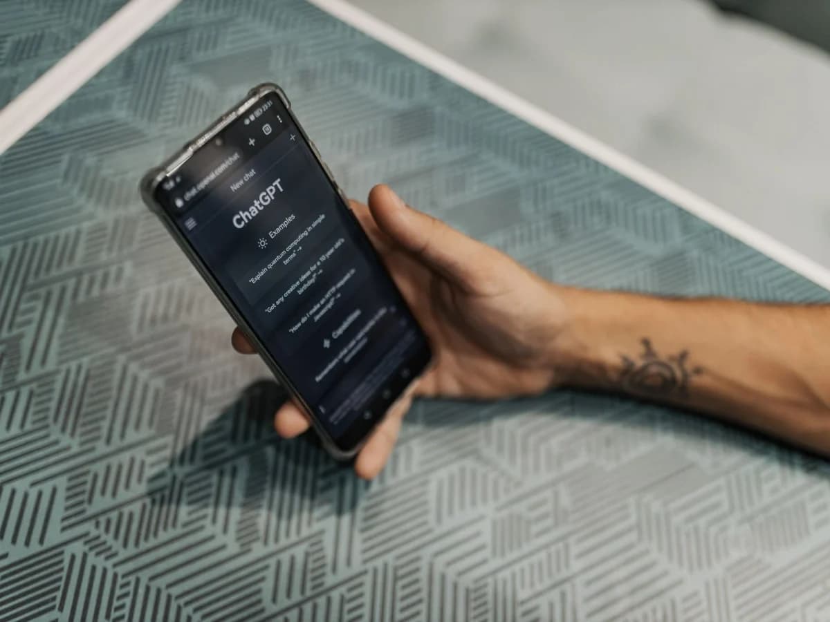 OpenAI GPT-6: Mão com tatuagem segurando um smartphone com a tela escura do ChatGPT, sobre uma superfície com padrão geométrico verde-acinzentado