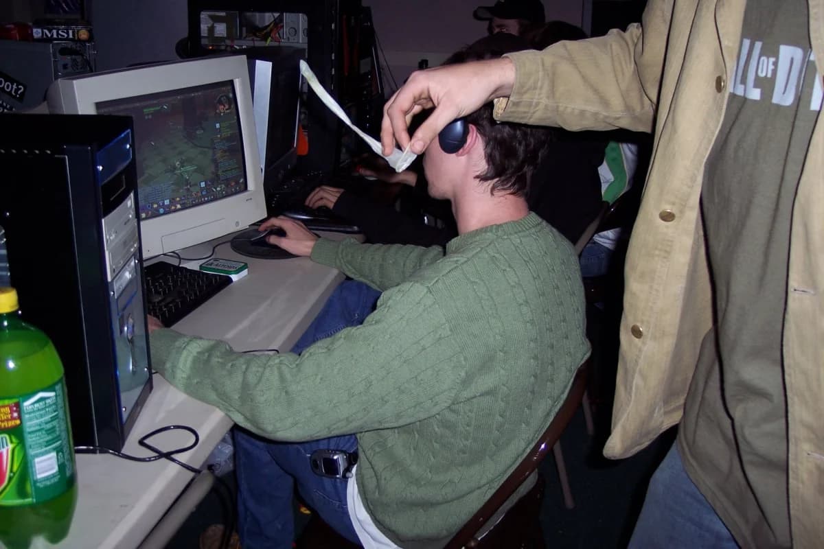 Lan house anos 2000, cybercafé com computadores de mesa e monitores CRT: Rapaz de suéter verde jogando em computador de gabinete com monitor CRT, enquanto outra