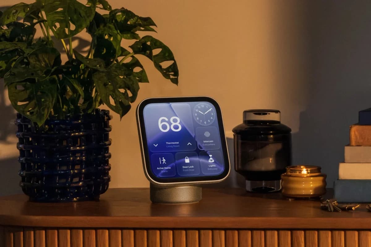 Apple HomeView: Display quadrado com tela azul mostrando temperatura 68 e ícones de casa, sobre um aparador de madeira ao lado de uma planta e uma vela, em ambi
