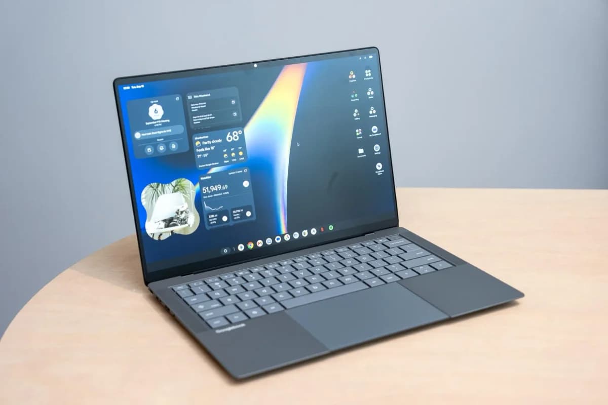 Google Googlebook: Notebook cinza-escuro aberto sobre mesa de madeira, em ângulo diagonal, com a tela exibindo widgets e ícones na área de trabalho
