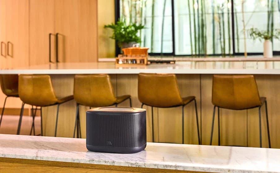 JBL Cove: Caixa de som preta retangular sobre um balcão de mármore, com banquetas de couro e vidro com bambu ao fundo.