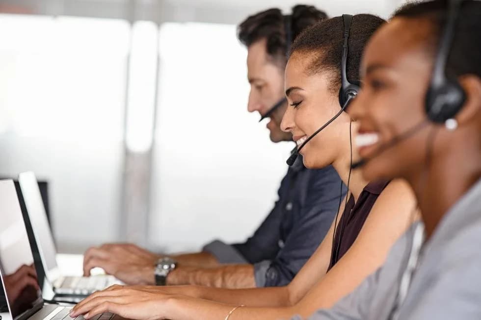 Serviço de Atendimento ao Consumidor (SAC): Três atendentes de perfil com fones de ouvido trabalhando em notebooks, sorrindo, em ambiente claro