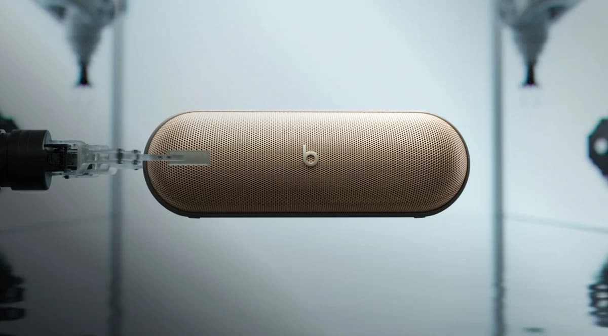 Beats Pill: Caixa de som dourada suspensa em fundo cinza-claro, de frente, com um bico transparente encostado na grade lateral.