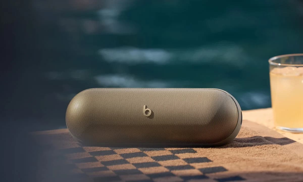 Beats Pill: Caixa de som dourada sobre uma toalha xadrez à beira de uma piscina, ao lado de um copo com bebida, em tons quentes.