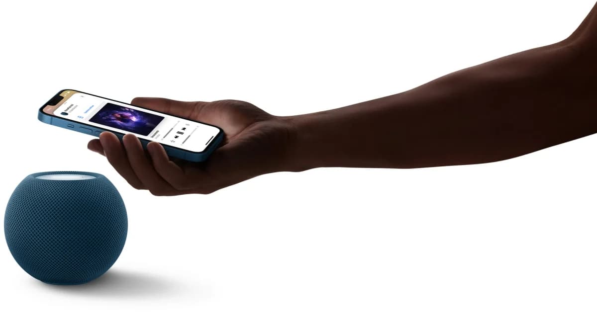 Apple HomePod mini: Caixa de som azul esférica no chão, ao lado de um braço e uma mão segurando um iPhone com tela de música, fundo branco.