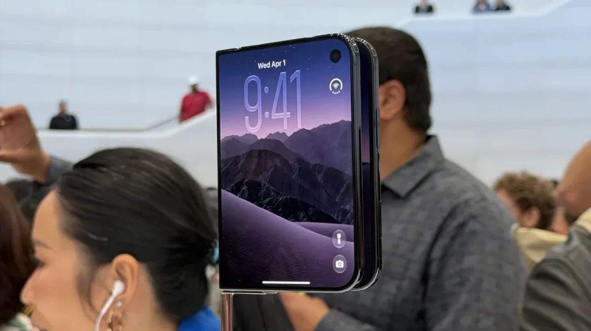 Apple iPhone Duo: Foto de evento mostrando celular dobrável em pé, com tela de bloqueio escura com montanhas, e pessoas ao redor em segundo plano.