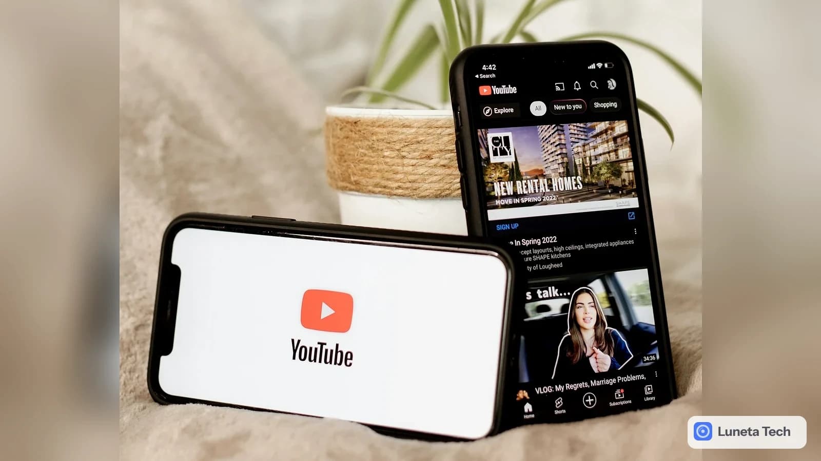 YouTube YouTube Music, selo de faixas feitas com IA: Dois celulares sobre tecido bege, um com logo do YouTube na tela branca e outro com feed de vídeos em tela