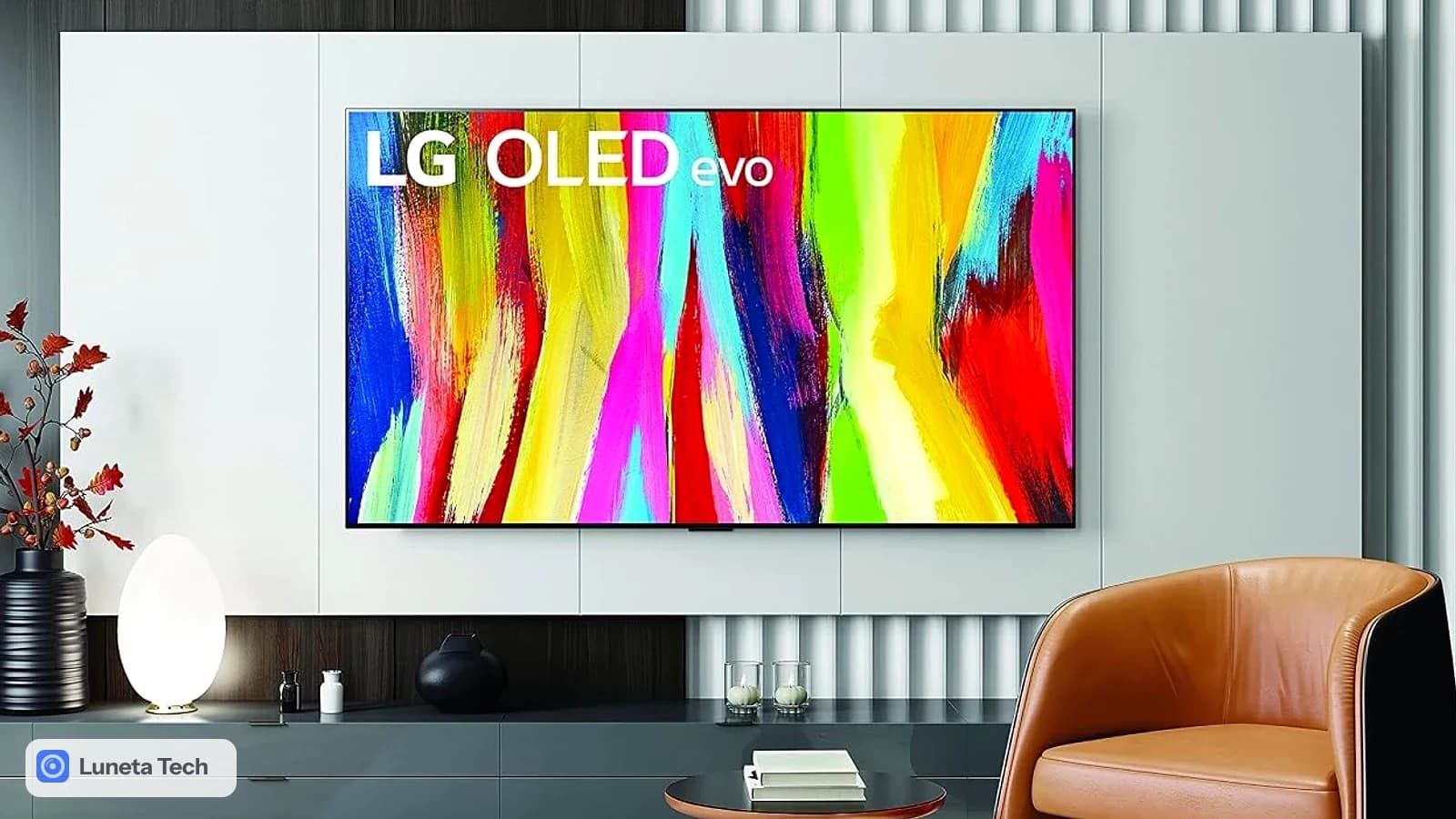 LG OLED evo C5: TV OLED grande montada na parede de sala clara, tela com pintura colorida em traços, poltrona de couro caramelo ao lado.
