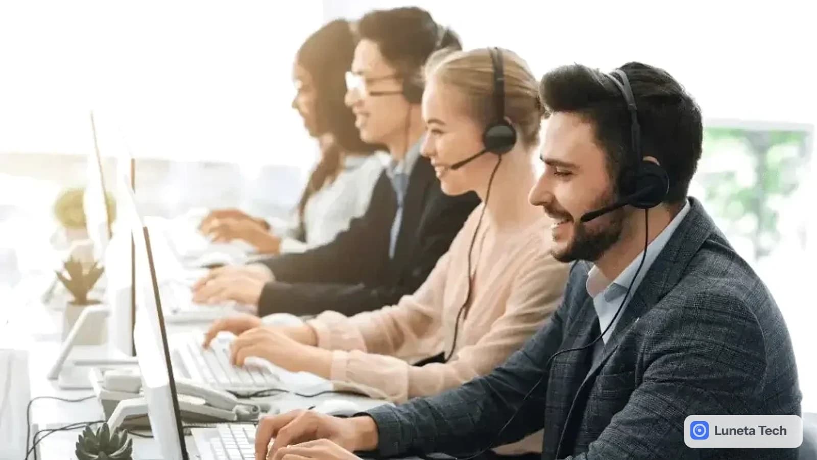 Serviço de Atendimento ao Consumidor (SAC): Atendentes com headset sorrindo diante de computadores em fileira, com luz clara ao fundo