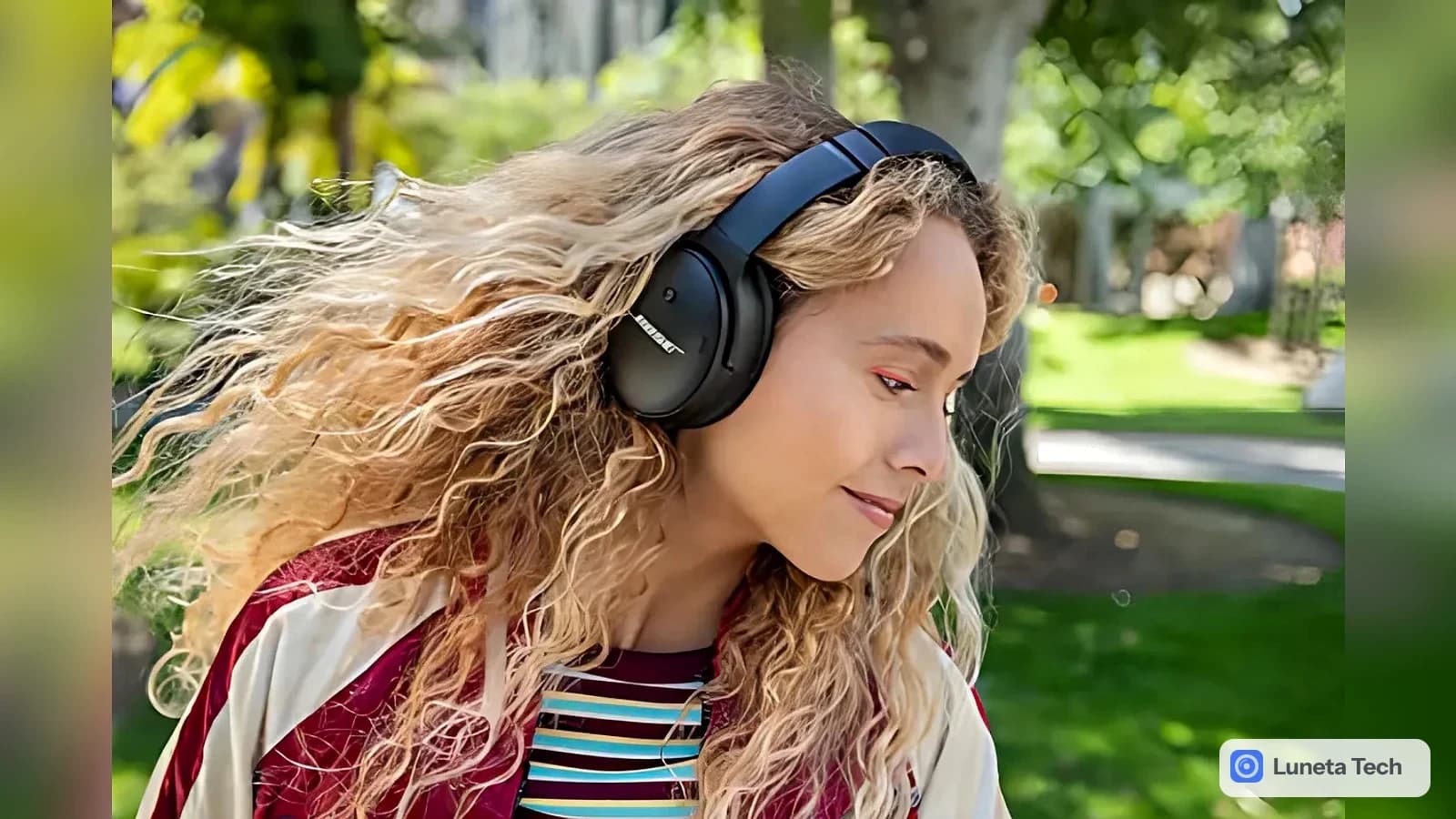 Bose QuietComfort Headphones (2ª geração): Mulher de cabelo cacheado usando fone over-ear preto ao ar livre, em um parque com árvores ao fundo.