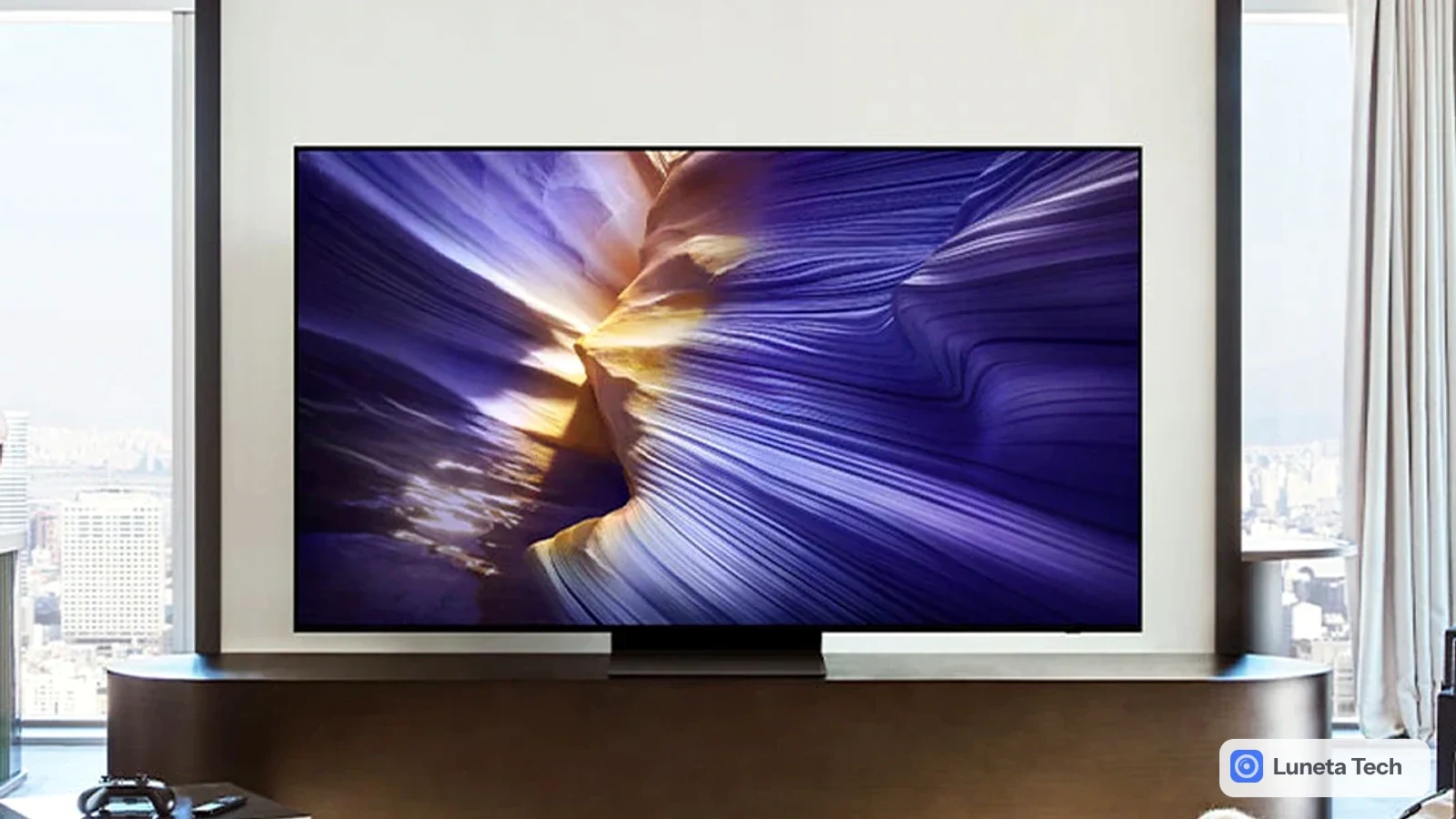 Samsung TV: Televisor OLED fino de frente sobre bancada escura em sala clara, com imagem roxa abstrata na tela, sofá de couro preto e manta à direita.