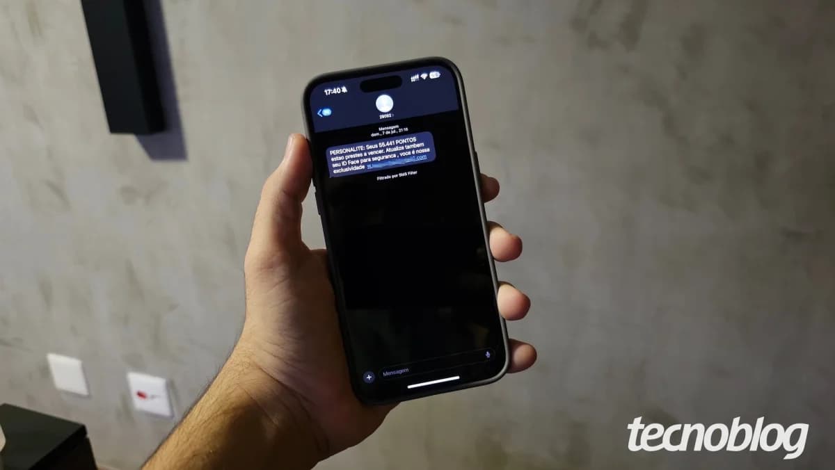 Phishing golpe de encomenda SMS: Mão segurando celular preto com mensagem de texto na tela escura, em ambiente interno com parede de concreto