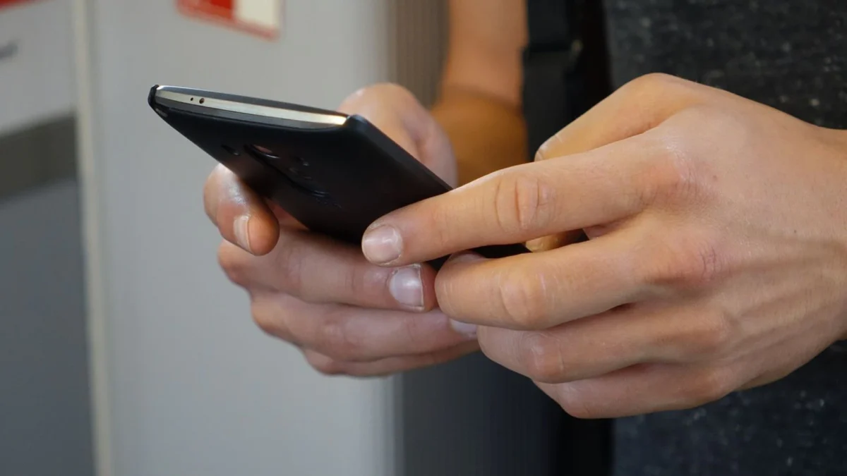 Phishing golpe de encomenda SMS: Duas mãos segurando e digitando em celular escuro, visto de lado, com fundo desfocado