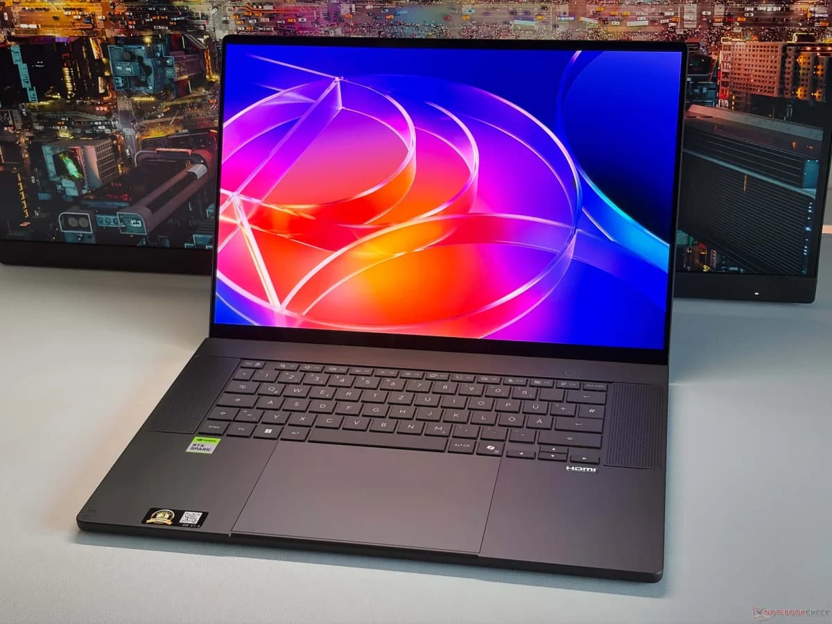 Asus ProArt P16: Notebook preto de frente, tela aberta exibindo arte colorida em tons de roxo, laranja e azul, sobre mesa cinza; ao fundo, monitor com cidade no