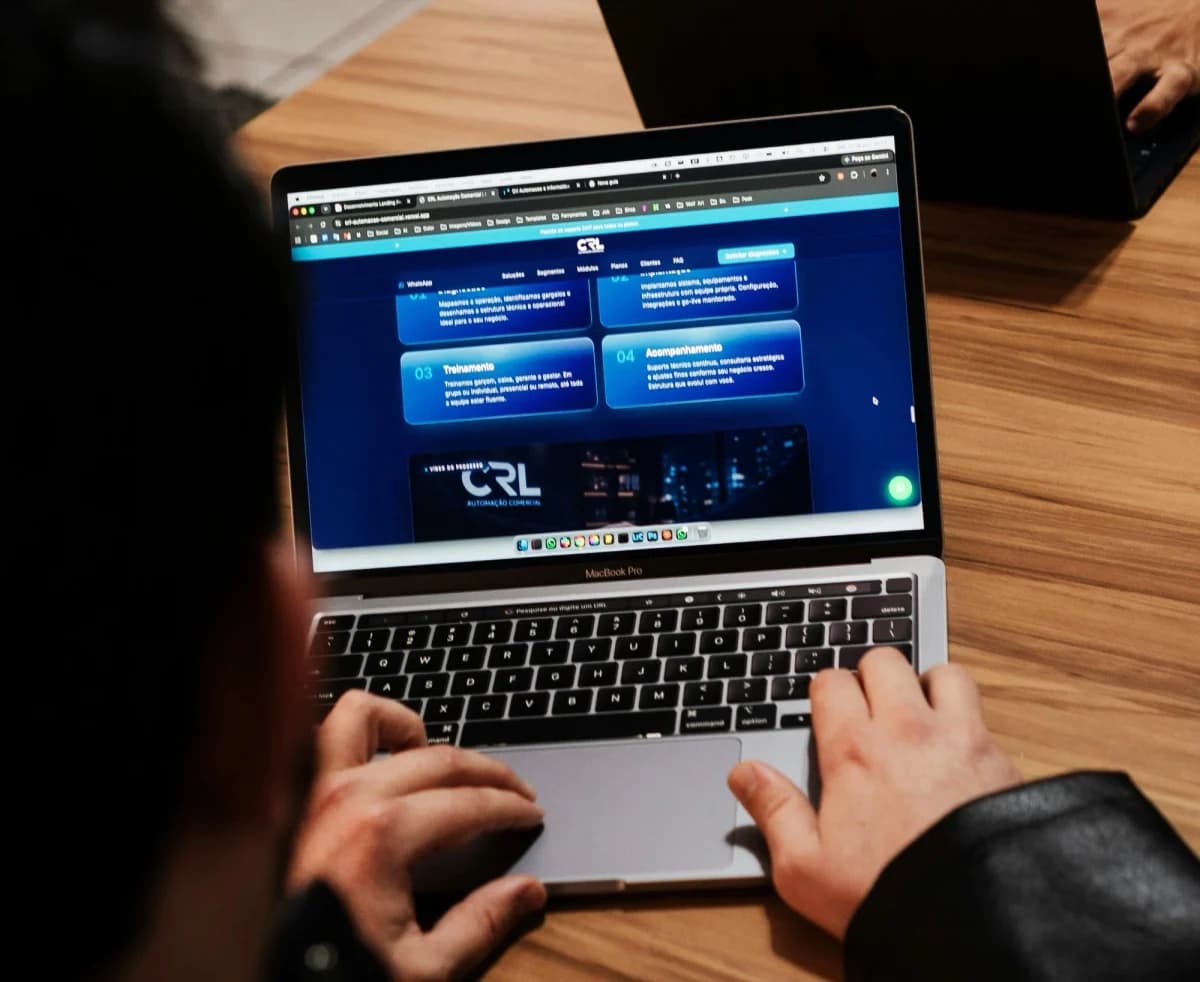 Apple MacBook Pro: Pessoa de costas usando notebook prateado com tela mostrando um site azul, mãos sobre o teclado e o trackpad, em mesa de madeira.