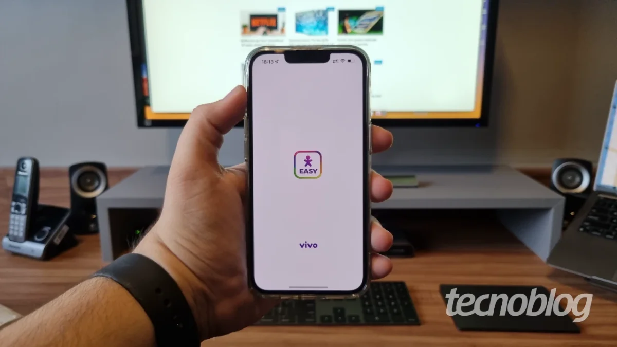 Vivo Vivo Travel, plano de roaming internacional da Vivo: Foto de mão segurando iPhone preto com a tela do aplicativo Easy da Vivo, com mesa, computador e telef