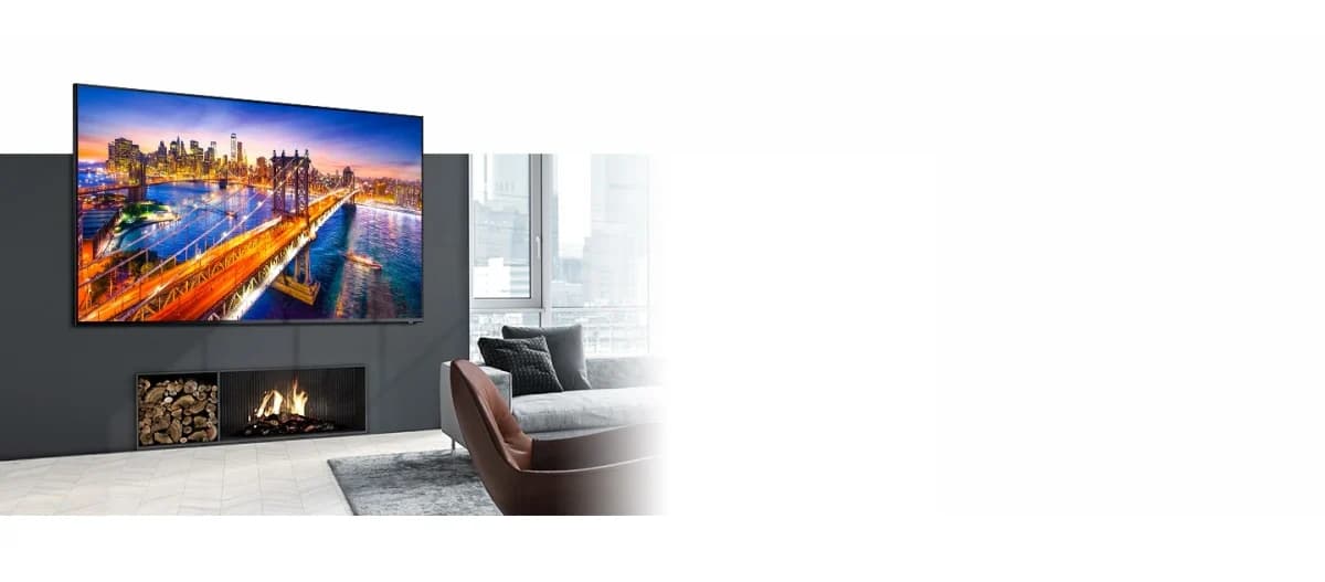 Samsung TV: Televisor preto fixado na parede de uma sala, com imagem de skyline noturno na tela, lareira ao lado e sofá marrom à direita.