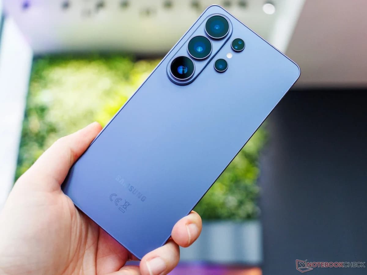 Samsung Galaxy S26 Ultra: Mão segurando smartphone azul-arroxeado de costas, mostrando o conjunto de câmeras traseiras e o logotipo Samsung em um ambiente exter