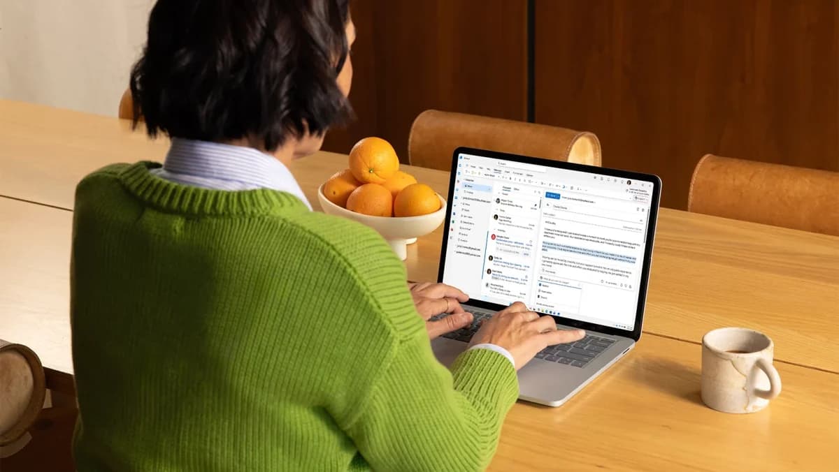 Microsoft Microsoft 365 Family: Mulher de costas, suéter verde, digitando em notebook prateado com e-mail aberto sobre mesa de madeira, ao lado de laranjas e um